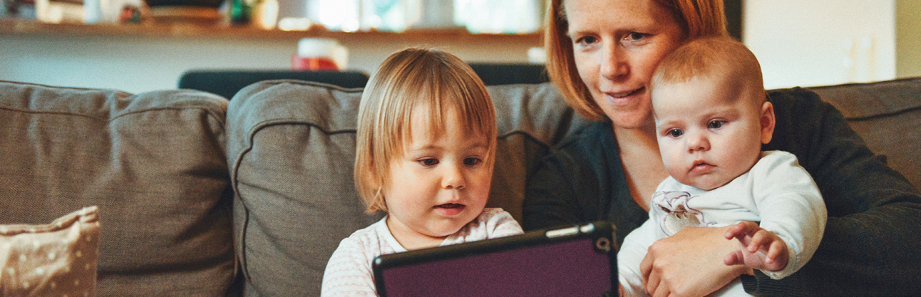 A mother and her two children sitting on a couch in front of a laptop enjoying Story Time virtually.